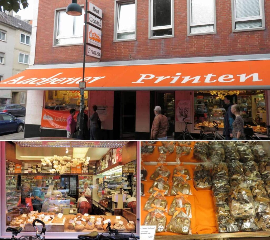 A bakery storefront in Aachen with orange signage reading "Aachener Printen." The window showcases various baked goods, reminiscent of Charlemagne's era, while the interior reveals customers and shelves brimming with products. Below, individually wrapped pastries rest on an orange cloth.