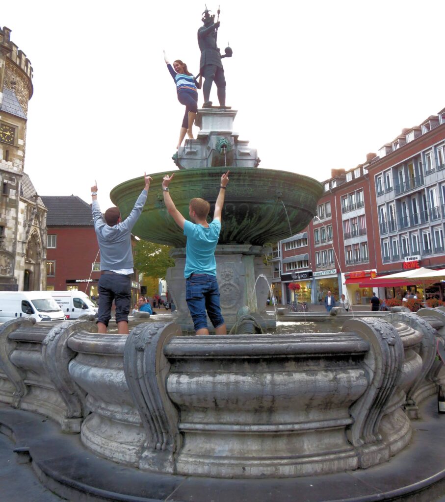 Three people are standing on a fountain in a city square in Aachen. Two men are on the lower level, pointing up, while a woman stands on the upper tier, posing with the statue of Charlemagne. Historic buildings and a few market stalls are visible in the background of this German scene.