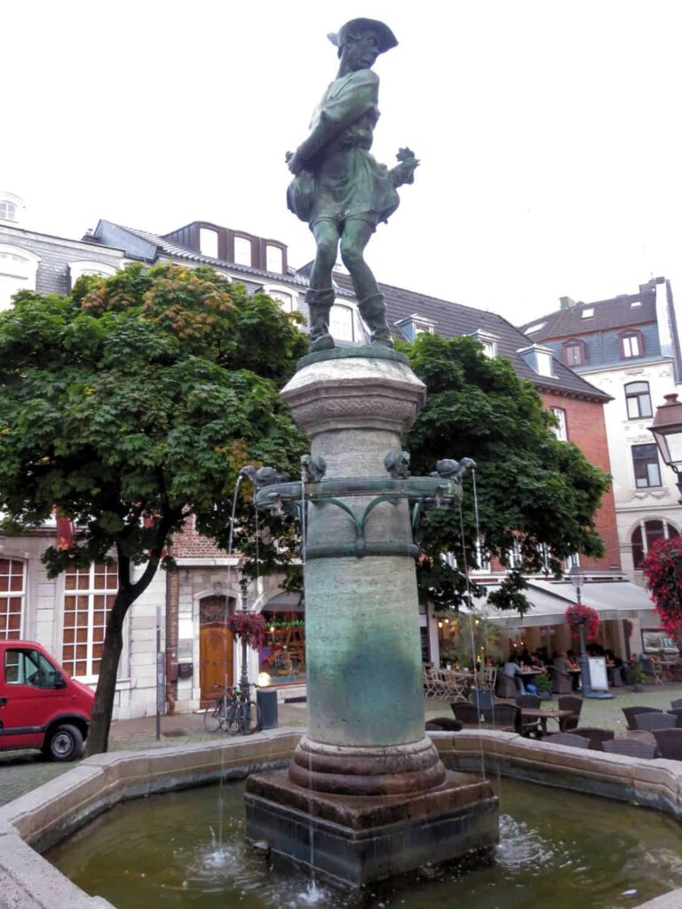 A bronze statue of Charlemagne stands atop a decorative pedestal in the center of a fountain in Aachen, Germany, surrounded by trees and buildings. Water flows from four spouts into the fountain below. A red car is parked nearby, and a café is in the background.