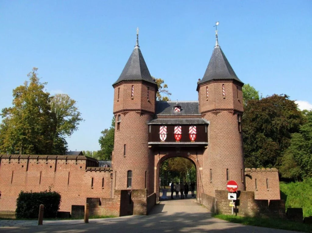 A brick castle gate, reminiscent of De Haar Castle, boasts two round towers and a central archway. Adorned with decorative shields and a red sign at the entrance, the grand structure evokes tales of the Knights of the Round Table. Trees frame it under a clear blue sky.