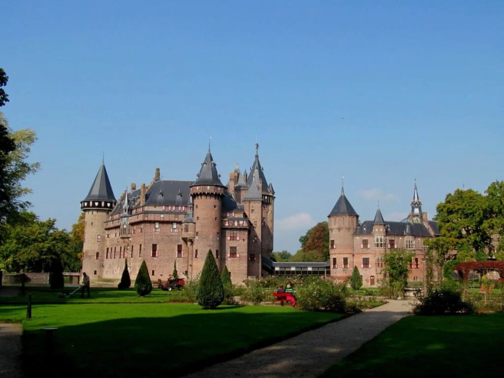 De Haar Castle, reminiscent of the era of the Knights of the Round Table, stands with its tall, pointed towers and red-brick walls amidst a landscaped garden with lush green lawns and winding pathways under a clear blue sky.