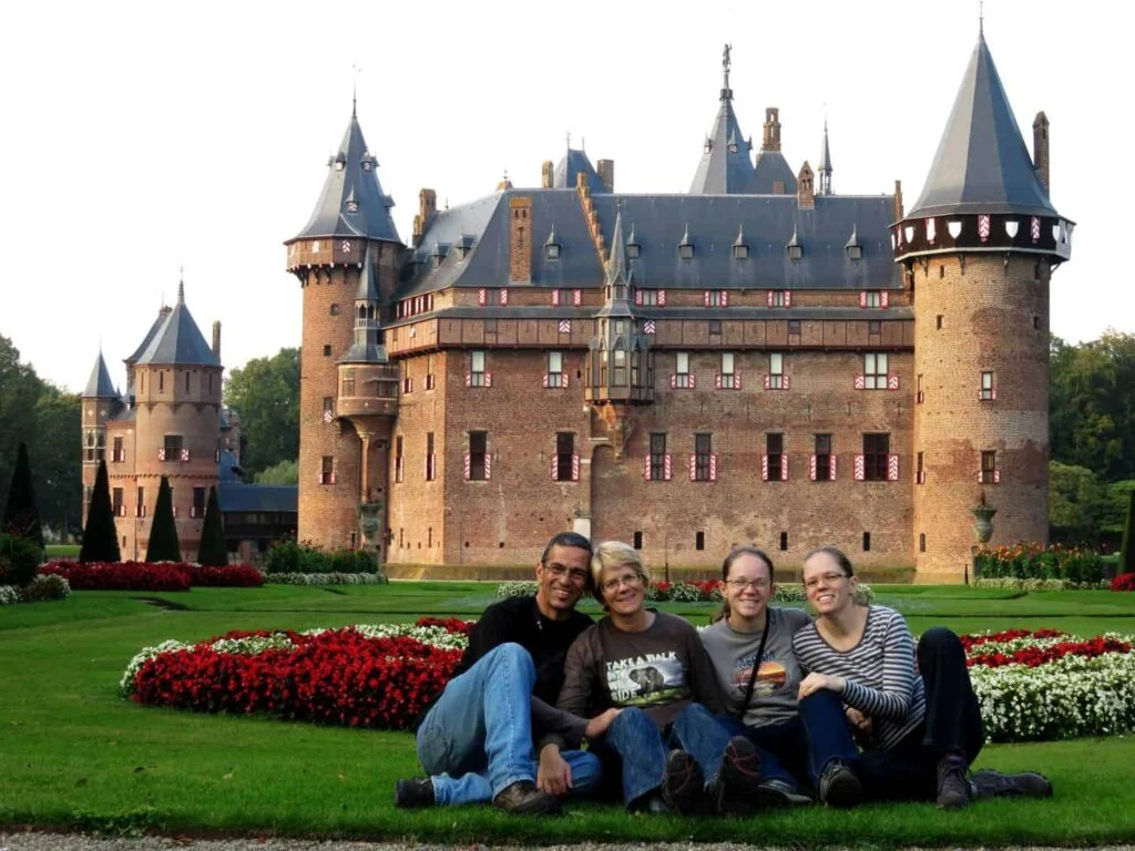Four people sit on the grass in front of the majestic De Haar Castle, its turrets reminiscent of the Knights of the Round Table era. On this bright day, red and white flowers adorn the green lawn, evoking an enchanting sense of history. All four appear relaxed and happy.