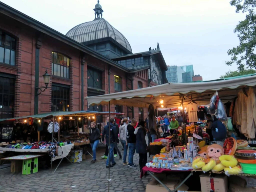 Amid the bustling ambiance of Hamburg's Fischmarkt, people browse stalls brimming with colorful toys and clothing. A historic brick building with a domed roof stands in the background, under an overcast sky that adds a cozy touch to the vibrant market scene.