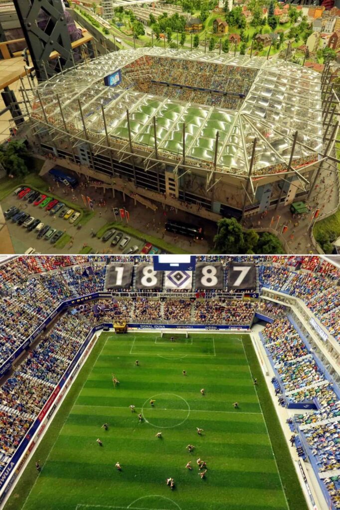 Aerial view of a detailed scale model at Miniatur Wunderland in Hamburg, Germany, showcasing a packed soccer stadium complete with a roof and surrounding parking lot. The model captures an ongoing match with players on a green field and stands brimming with vibrant spectators.