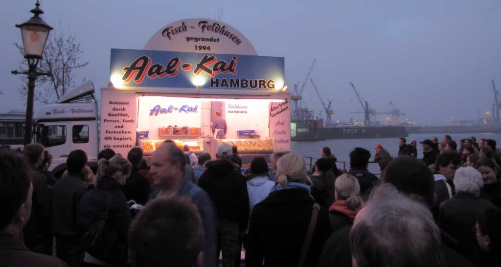 A crowd gathers at the bustling Fischmarkt in front of a brightly lit seafood stall labeled "Aal-Kai Hamburg" near the harbor. The evening sky is overcast, and cranes are silhouetted in the background as people eagerly queue for food.